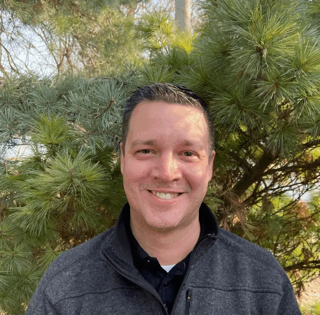 Man smiling in front of pine tree.
